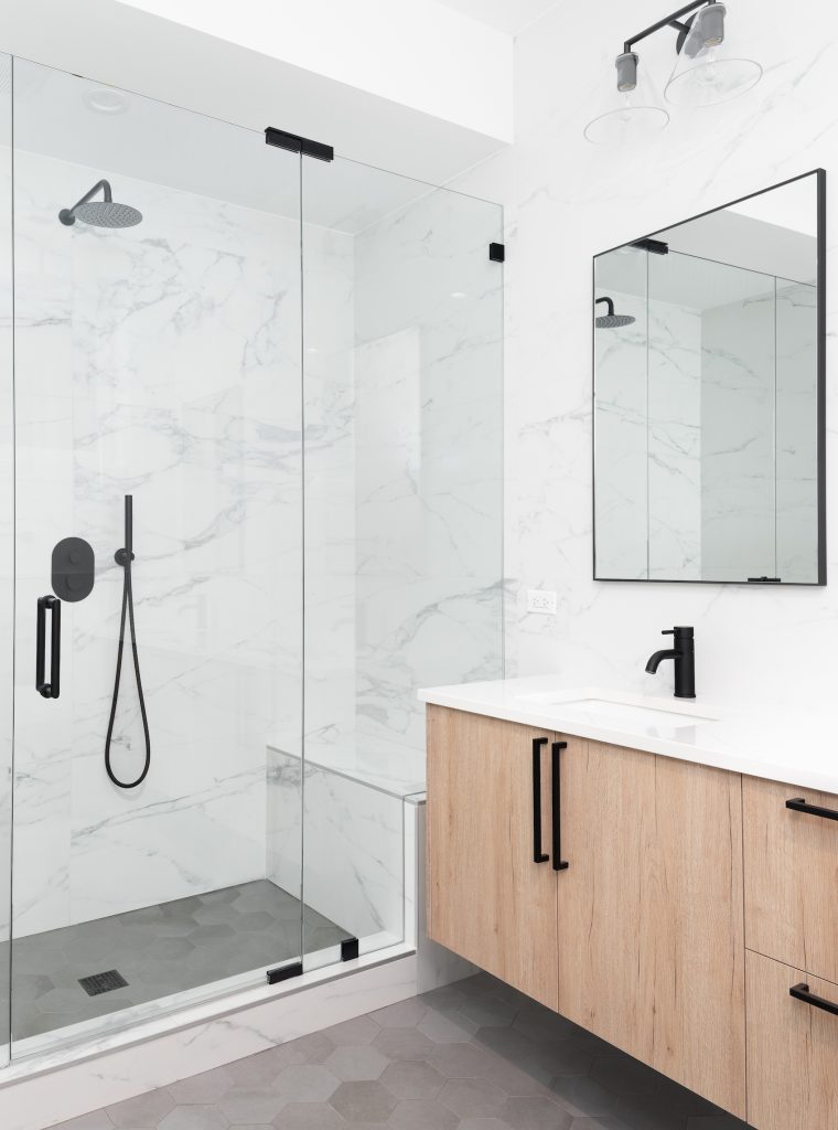 A beautiful luxury, modern bathroom with a light wood cabinet, walk-in shower with marble tiled walls, and black faucets and hardware.